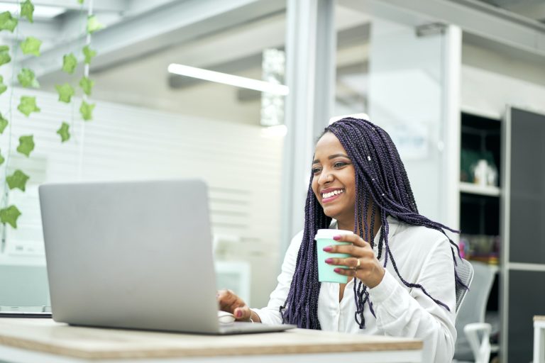 Smiling businesswoman sitting at office desk holding coffee while working on laptop.